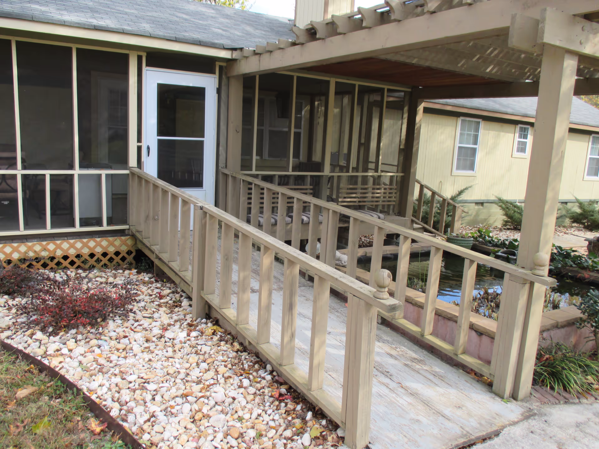A wooden ramp with railings leading to a screened porch entrance of a building. The porch has a white door and is surrounded by a garden area with rocks and plants. Another building with windows is visible in the background.