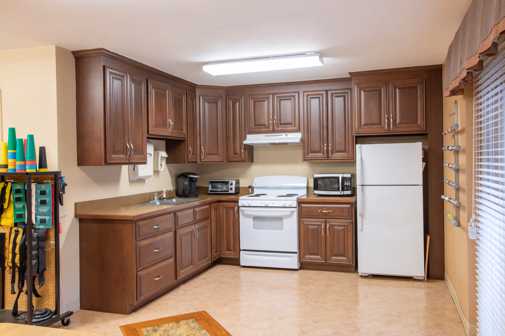A kitchen area with wooden cabinets, a white stove, a microwave, a toaster oven, a coffee maker, and a white refrigerator. There is a sink with a paper towel dispenser above it. On the left side, there is a rack holding colorful exercise equipment. On the right side, there is a window with blinds and a wall-mounted rack holding rolled-up items.