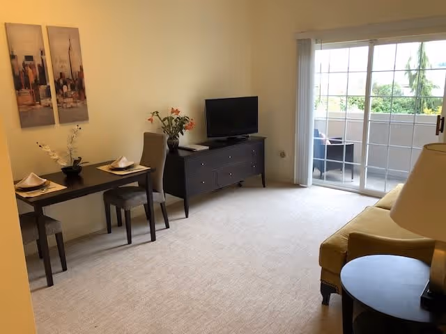 A bright living room with beige carpet and light yellow walls. There is a small dining table with two chairs on the left side, set with plates and napkins. A black TV stand with a flat-screen TV and a vase with flowers is against the wall. On the right side, there is a yellow sofa and a round side table with a lamp. Large sliding glass doors lead to a balcony with outdoor seating and greenery visible outside.