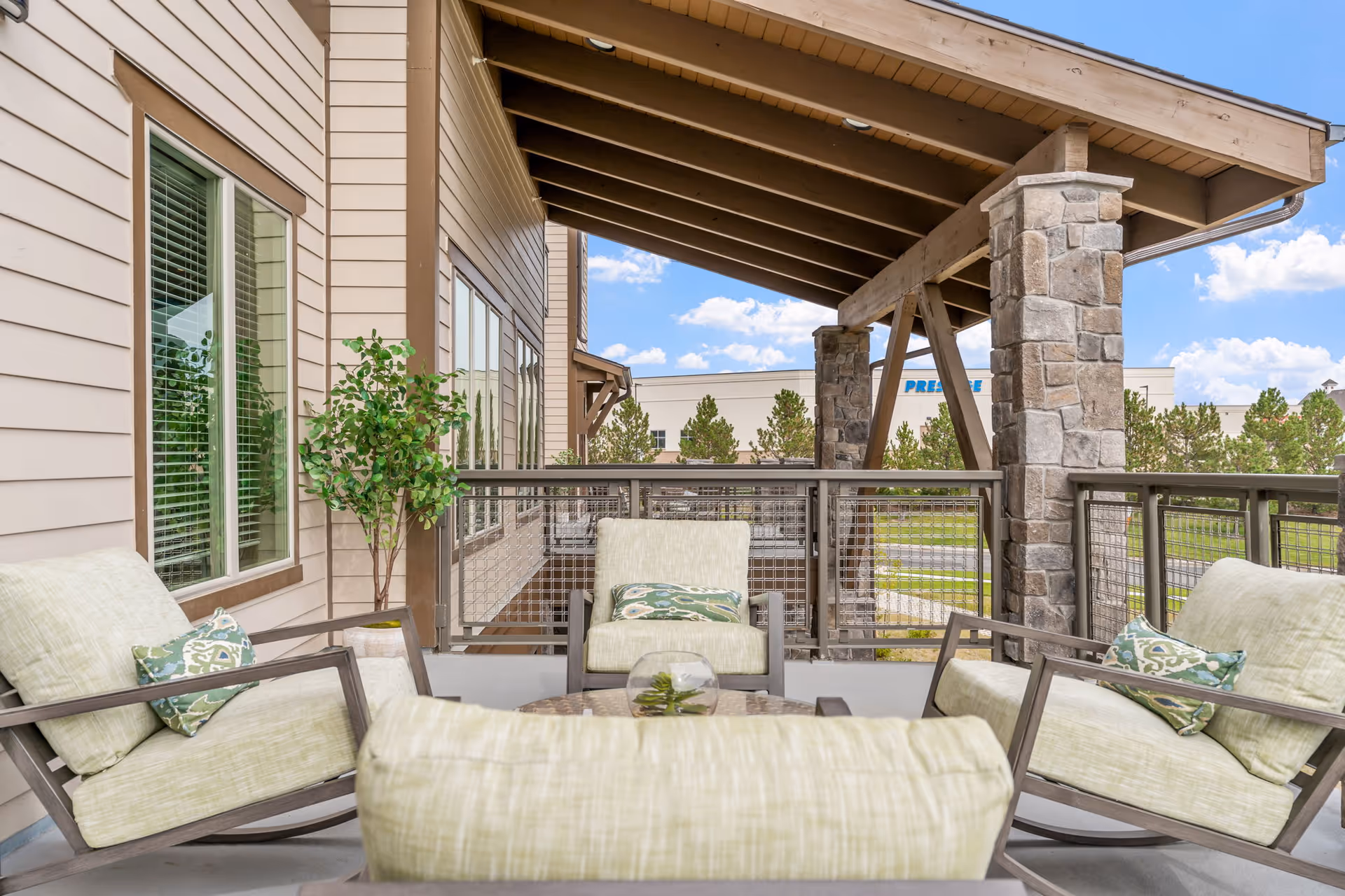 Outdoor covered patio area with cushioned chairs and a small table, surrounded by a railing and stone pillars, with a view of trees and a building in the background under a blue sky with clouds.