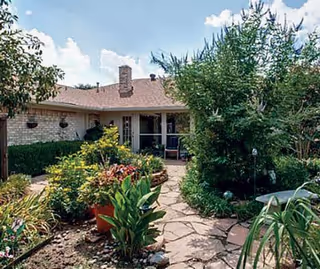 A garden pathway leading to the entrance of a single-story building with a chimney. The pathway is surrounded by lush green plants, shrubs, and flowers under a partly cloudy sky.