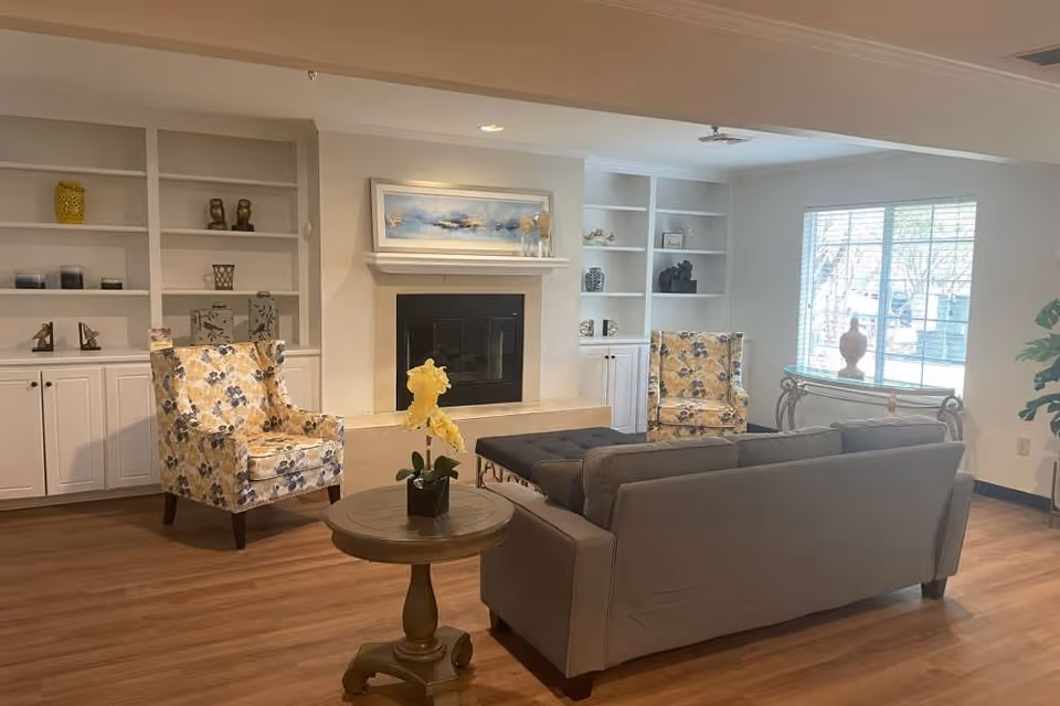 A cozy living room with a gray sofa, two floral armchairs, a round wooden side table with a yellow orchid plant, a fireplace with a painting above it, built-in white shelves with decorative items, and a large window letting in natural light.