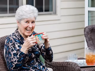 An elderly woman with short white hair sitting outdoors on a wicker chair, smiling and holding a colorful mug. There is a glass of iced tea and a glass of water on a table next to her, with the exterior siding of a building in the background.