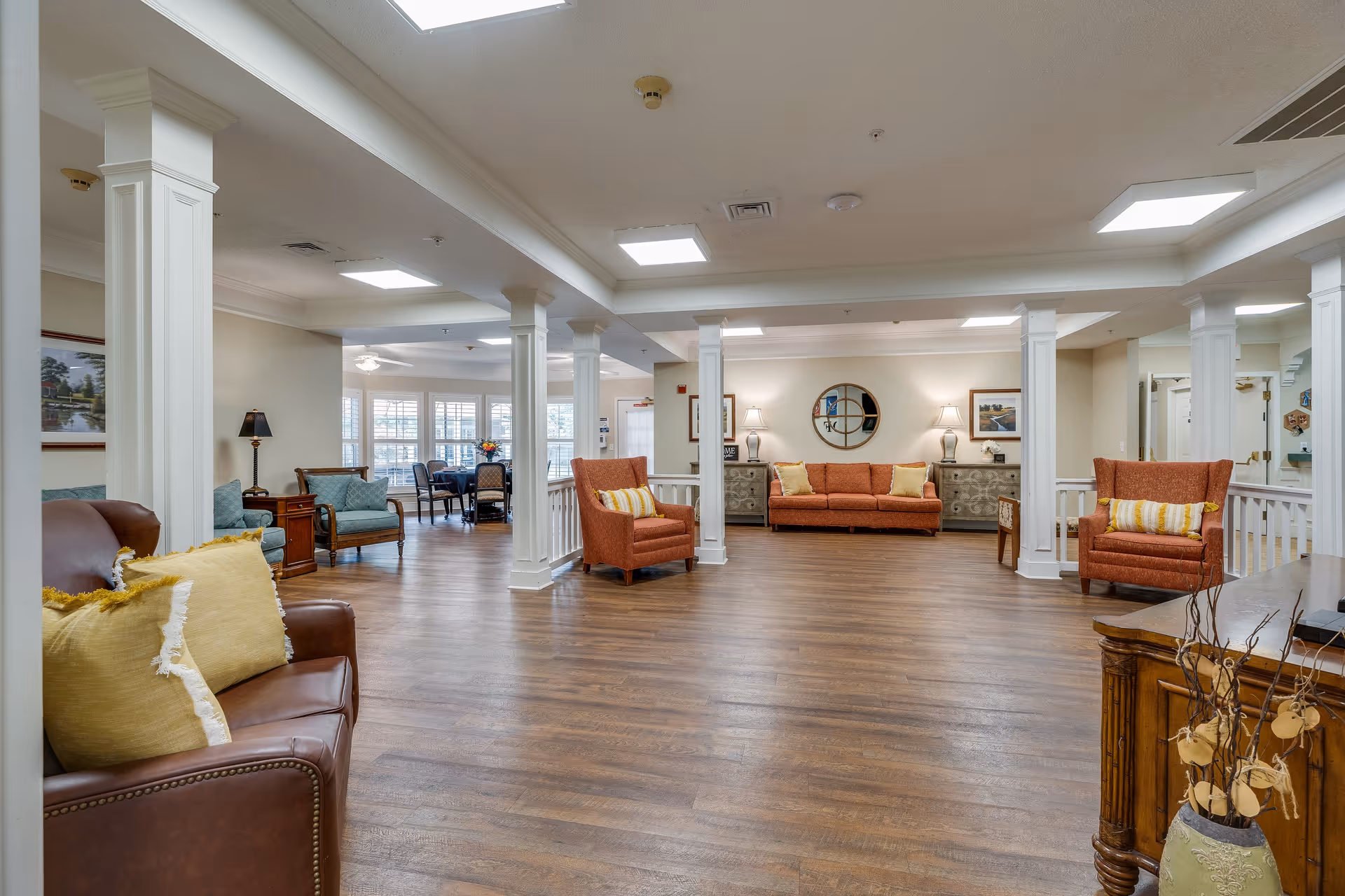 A spacious and well-lit living area in a senior living facility with wooden flooring, multiple seating options including brown leather and orange fabric armchairs and sofas with yellow cushions, white columns, and a dining area visible in the background near large windows with white blinds.