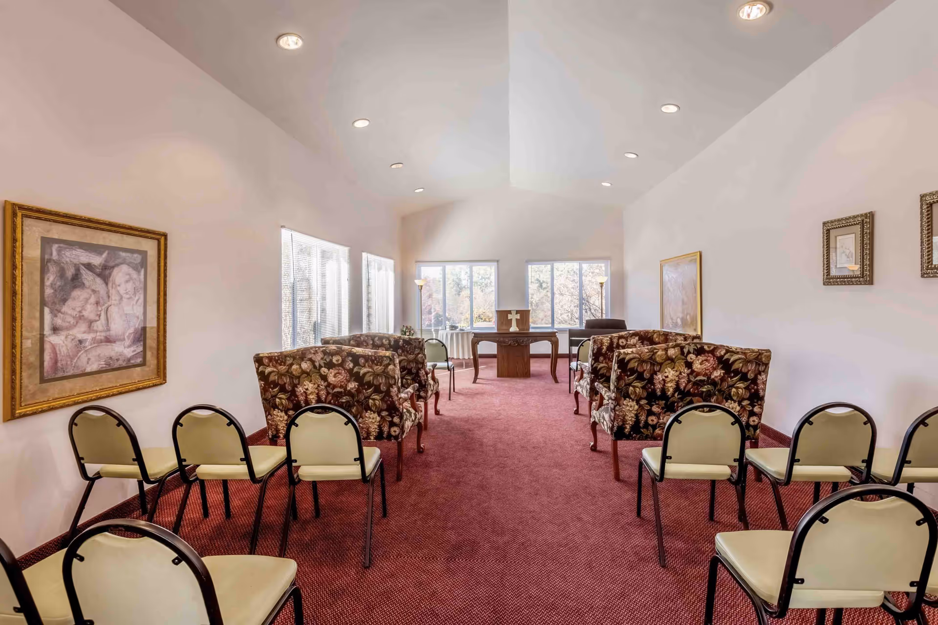 Interior community meeting/chapel room with rows of chairs and floral armchairs facing a small altar table under a high ceiling.