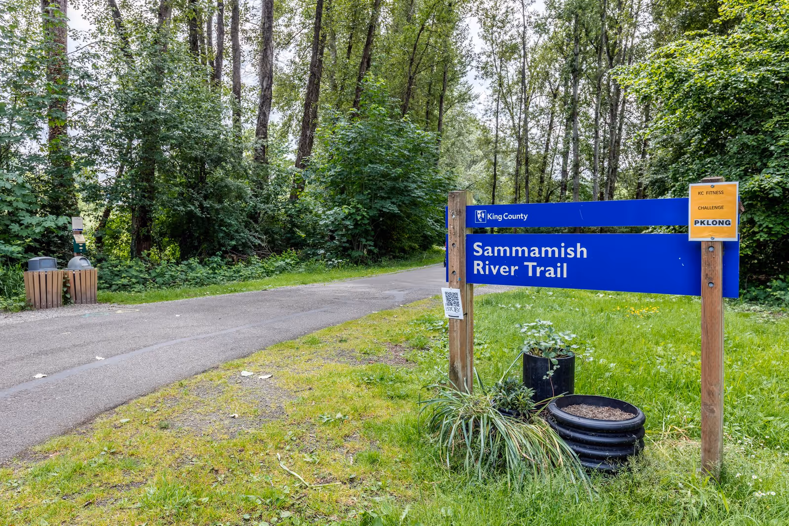A paved trail surrounded by lush green trees and grass with a blue sign that reads 'Sammamish River Trail' and a smaller yellow sign indicating a fitness challenge. There are two trash bins on the left side of the trail and some plants near the signpost.