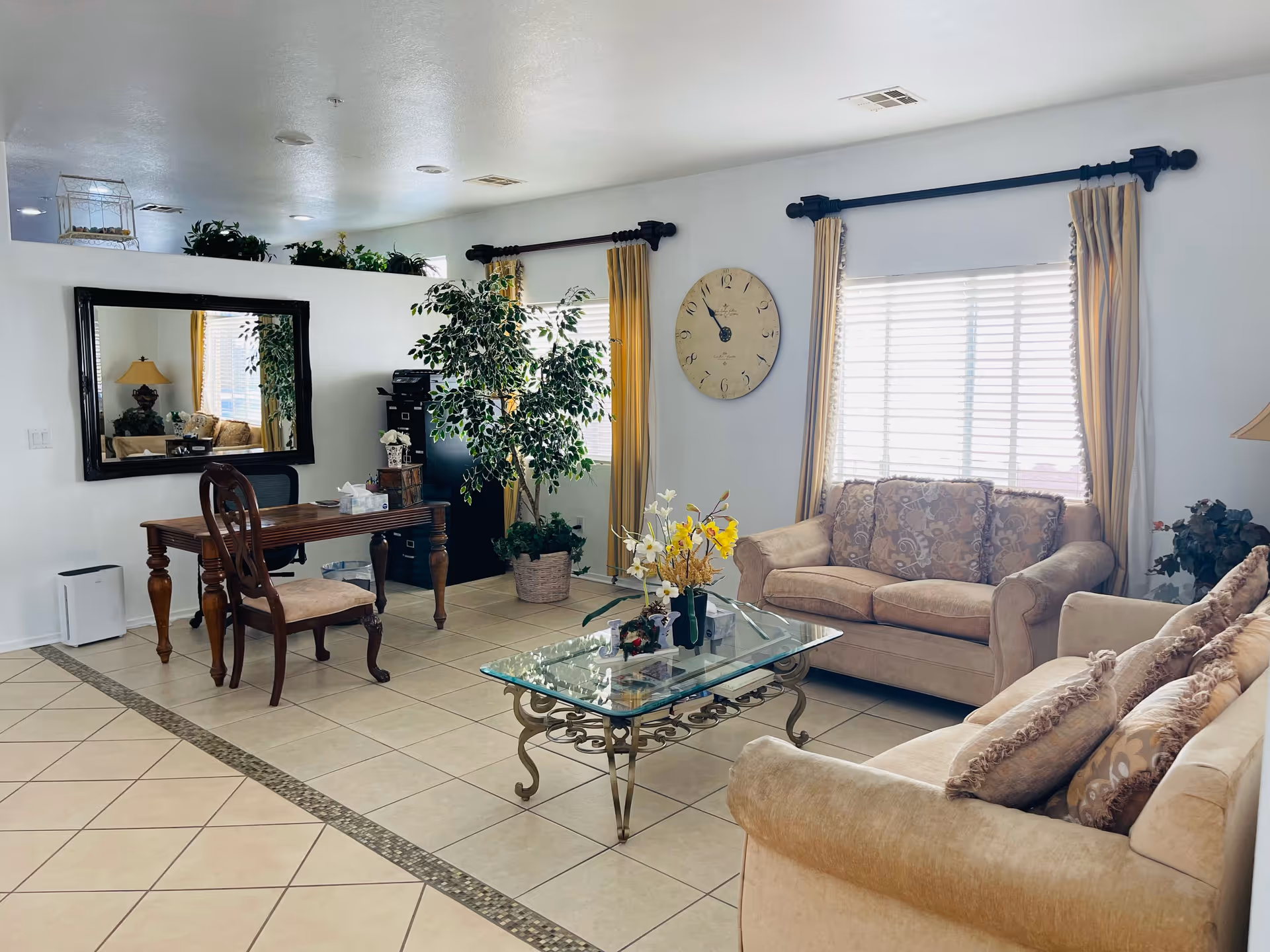 A bright living room with beige sofas adorned with patterned cushions, a glass coffee table with a floral arrangement, a wooden desk with a chair, a large wall clock, a tall potted plant, and a large mirror reflecting part of the room. The room has tiled floors and windows with yellow curtains.