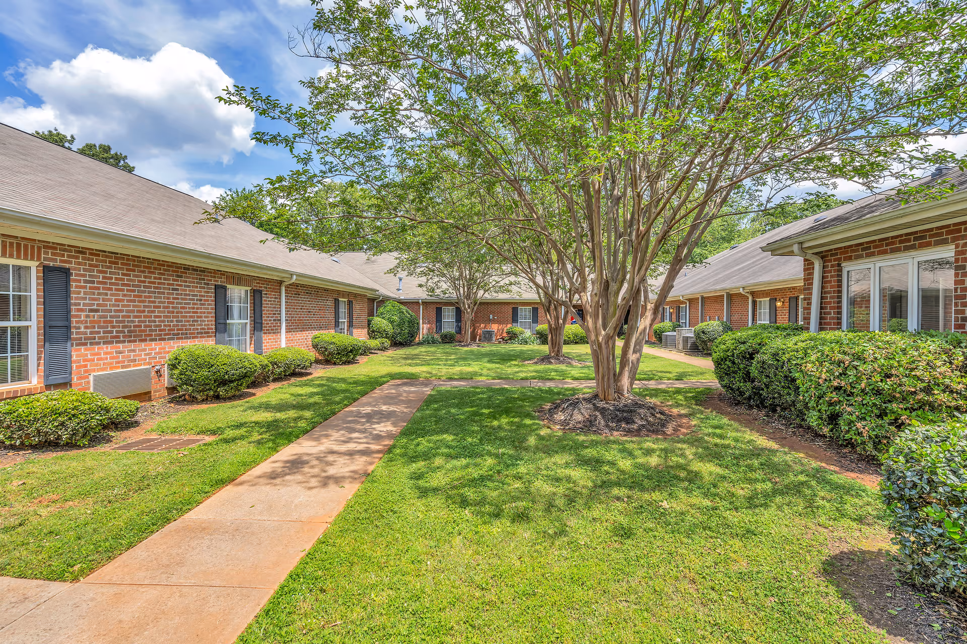 Outdoor courtyard area with a concrete walkway, green grass, bushes, and trees surrounded by single-story brick buildings with windows and shutters under a partly cloudy blue sky.