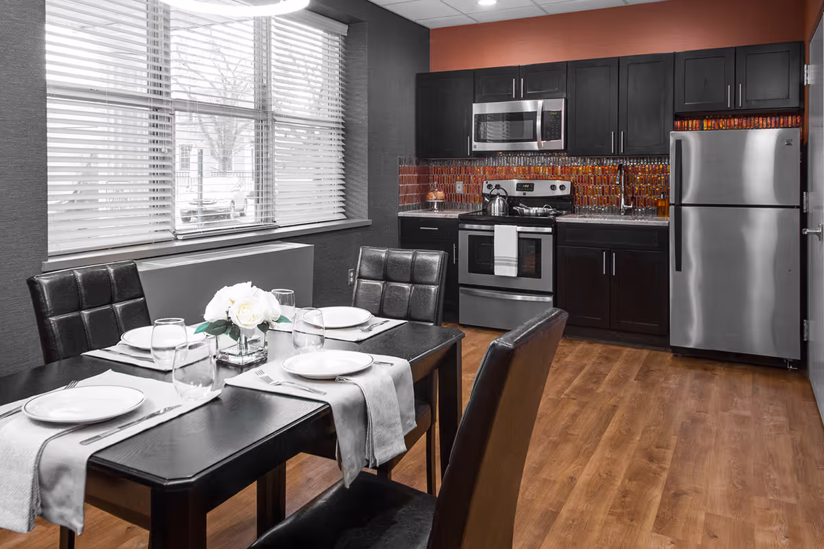 Modern dining area with a set table and chairs next to a kitchenette with dark cabinets, stainless steel appliances, and wood flooring.