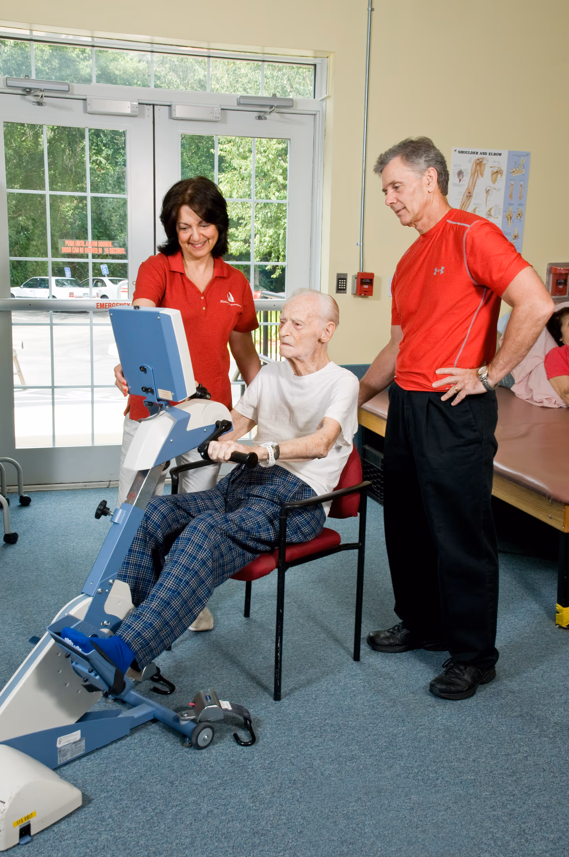 An elderly man seated on a chair using a physical therapy exercise machine while being assisted by a female therapist in a red polo shirt and a male therapist in a red athletic shirt in a room with large windows and exercise tables.