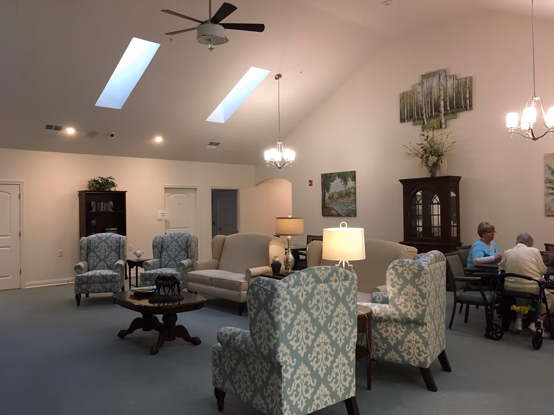 A bright assisted-living common room with patterned armchairs, sofas, lamps, skylights and two people seated at a table.