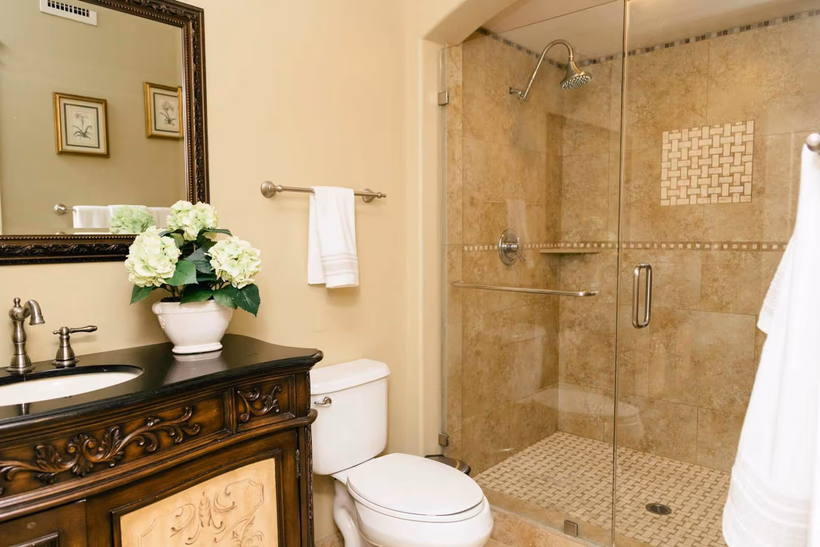 A bathroom with a decorative wooden vanity topped with a black countertop and a white sink. A potted plant with white flowers sits on the vanity. Above the vanity is a large framed mirror. Next to the vanity is a white toilet, and beside it is a glass-enclosed shower with beige tiles and a built-in bench. A white towel hangs on a towel rack on the wall.