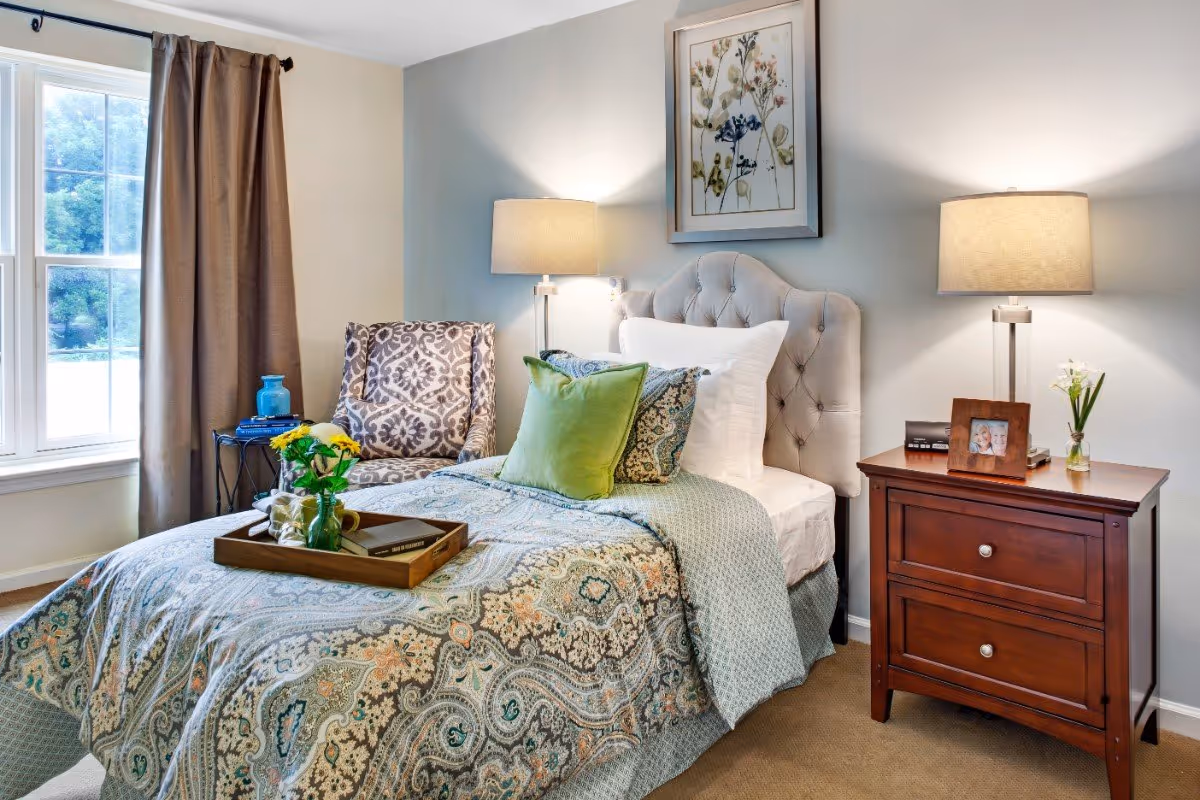 Well-decorated bedroom with an upholstered headboard, patterned bedding, nightstand with lamp and framed photo, and a window with curtains.