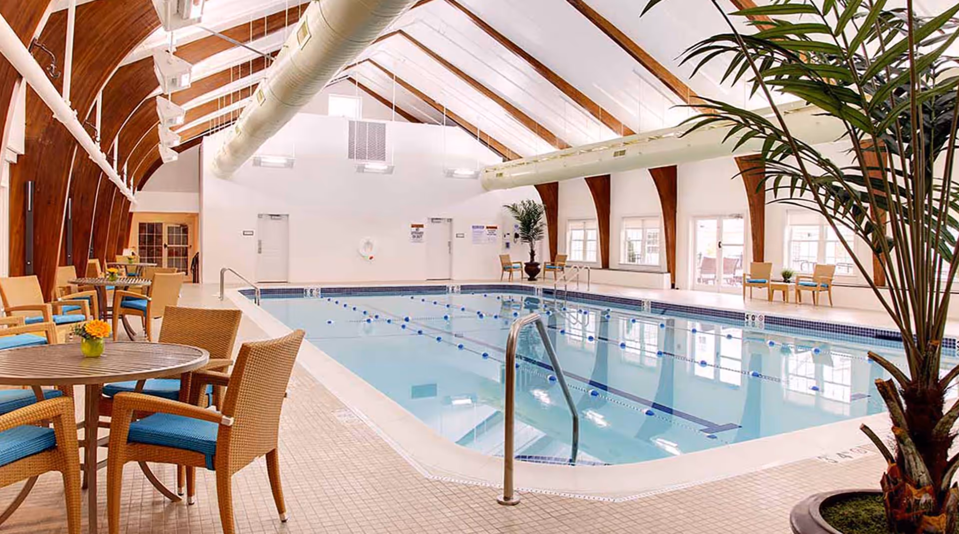 Indoor swimming pool with lane markers and seating tables under a vaulted wooden ceiling.