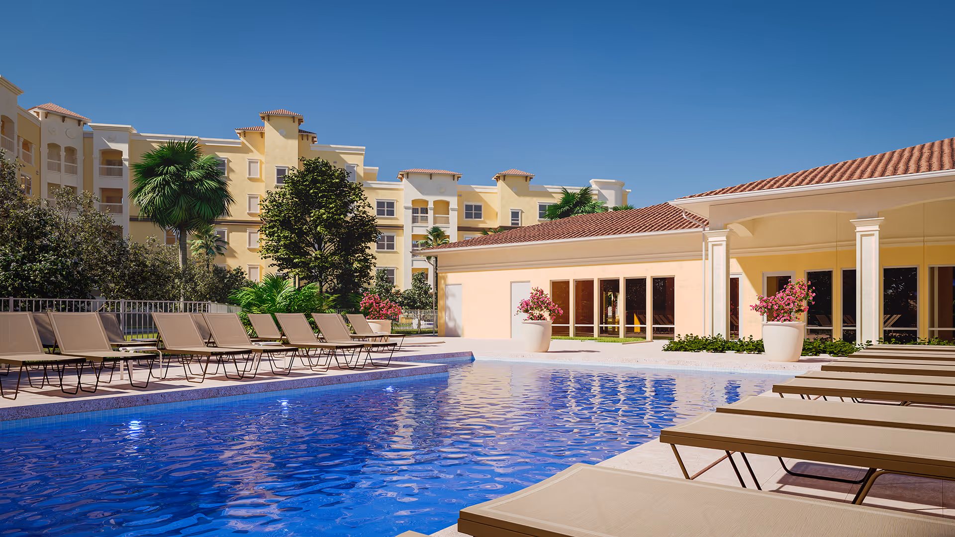 Outdoor swimming pool area with multiple beige lounge chairs arranged around the pool. The pool water is clear and blue, reflecting the bright sunny sky. In the background, there is a light yellow building with a red-tiled roof, large windows, and potted plants with pink flowers. Tall trees and a multi-story residential building are visible behind the pool area.