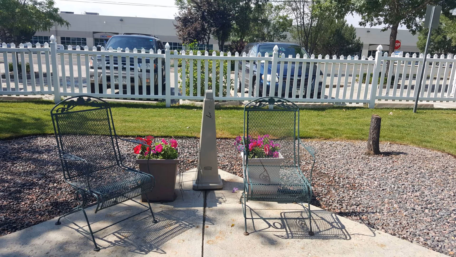 Two metal mesh chairs with armrests placed on a concrete patio area surrounded by gravel and grass. There are two flower pots with pink flowers between the chairs. A white picket fence and parked cars are visible in the background under a clear sky.
