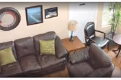 A cozy living room area featuring a brown leather sofa and matching armchair, both with green accent pillows. There is a wooden side table with a lamp between the sofa and armchair. On the wall above the sofa are three framed pictures, and a tall plant is placed near a window letting in natural light.