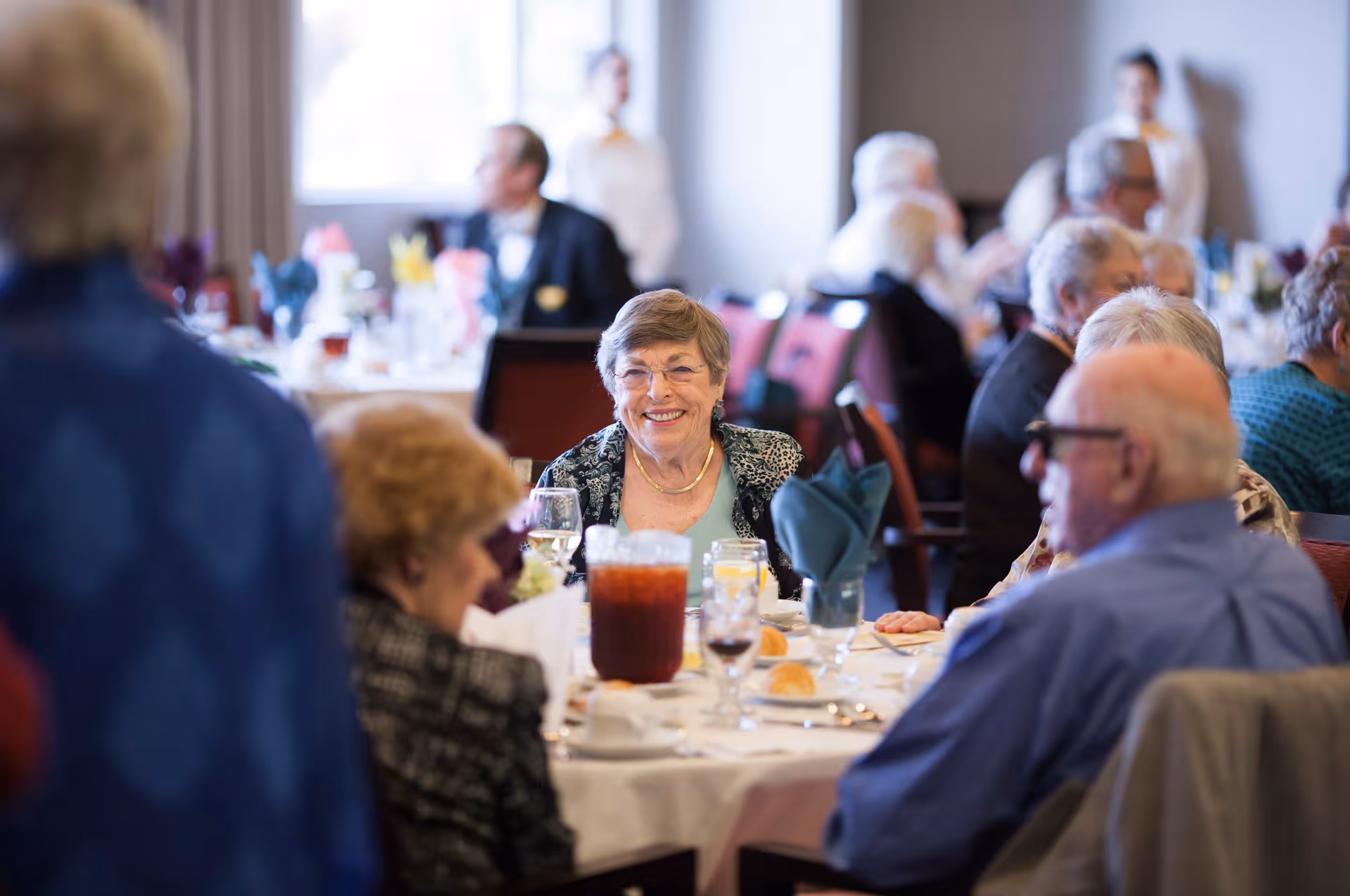 A group of elderly people seated around a dining table in a retirement community dining room, with plates, glasses, and a pitcher of iced tea on the table. One elderly woman in the center is smiling and looking towards the camera.
