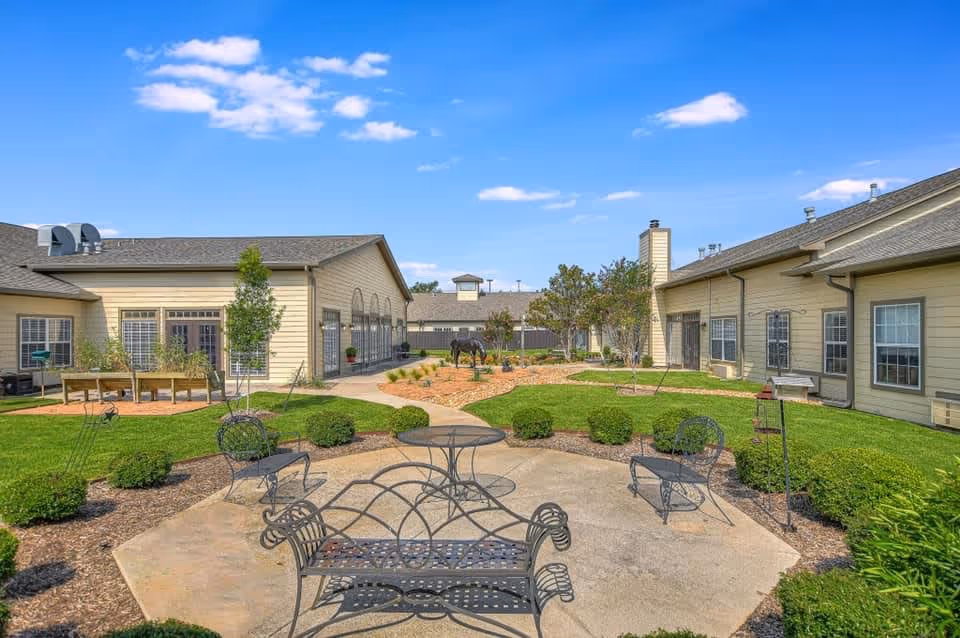 Outdoor courtyard area at Cedar Crest Senior Living of Lewisville featuring a paved patio with metal chairs and a table, surrounded by manicured bushes and green grass. Beige buildings with multiple windows and doors enclose the courtyard under a bright blue sky with scattered clouds.