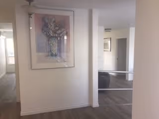 Interior view of a hallway in a senior living facility with light-colored walls and wood flooring. A framed floral painting is hung on the left wall. The hallway leads to other rooms with doors visible in the background.