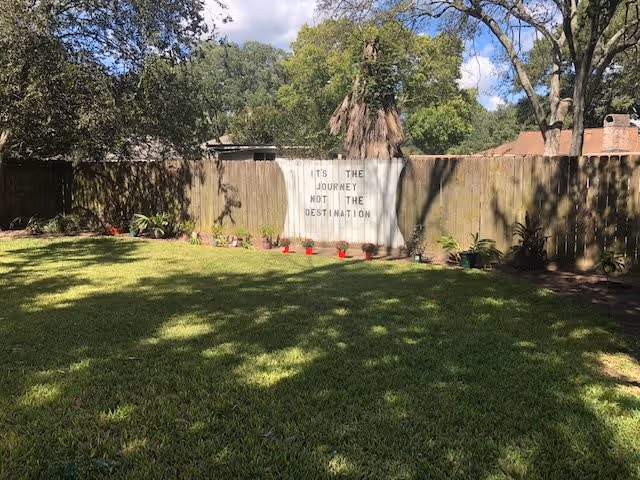 A backyard garden area with a well-maintained green lawn, bordered by a wooden fence. There are several small potted plants lined up against the fence. On the fence, a white board displays the quote 'IT'S THE JOURNEY NOT THE DESTINATION'. Trees and a partly cloudy sky are visible in the background.