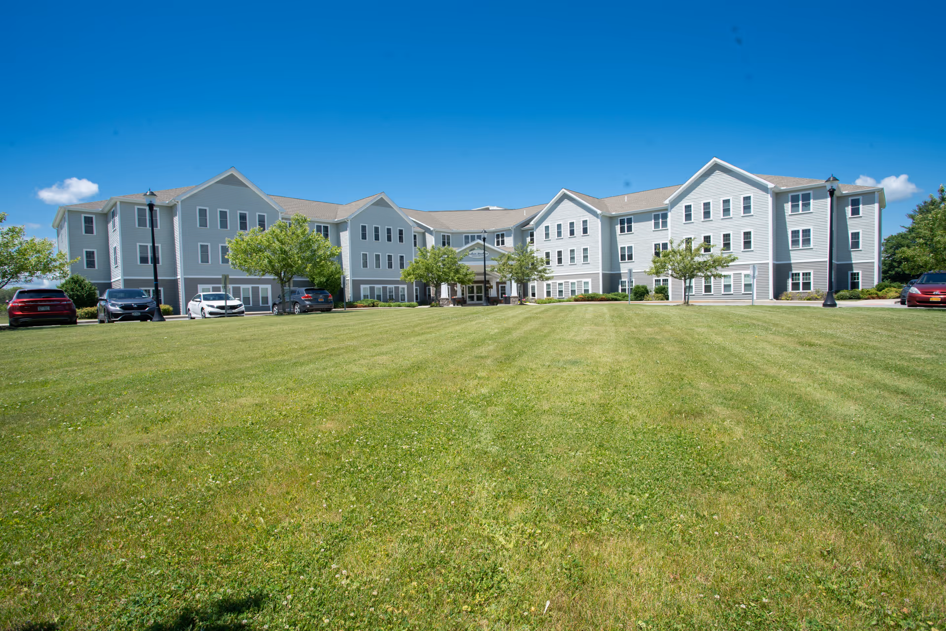 Three-story assisted living building viewed from a wide green lawn with cars parked along the driveway under a clear blue sky.