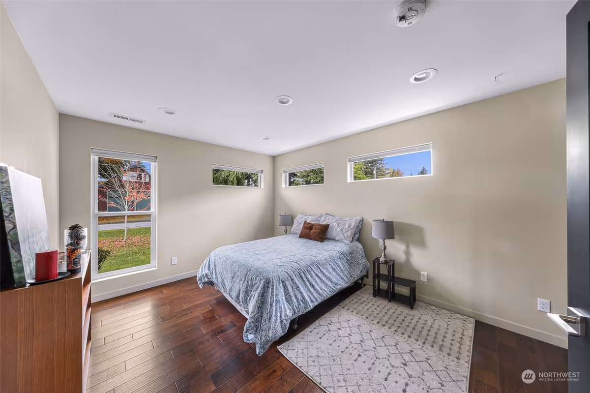 A bright bedroom with beige walls and dark wooden flooring. The room features a bed with a blue patterned comforter and two pillows, two bedside tables each with a lamp, a light-colored patterned rug, and three horizontal windows letting in natural light. There is also a tall window showing a view of a tree and a house outside. A wooden shelf with decorative items is visible on the left side.