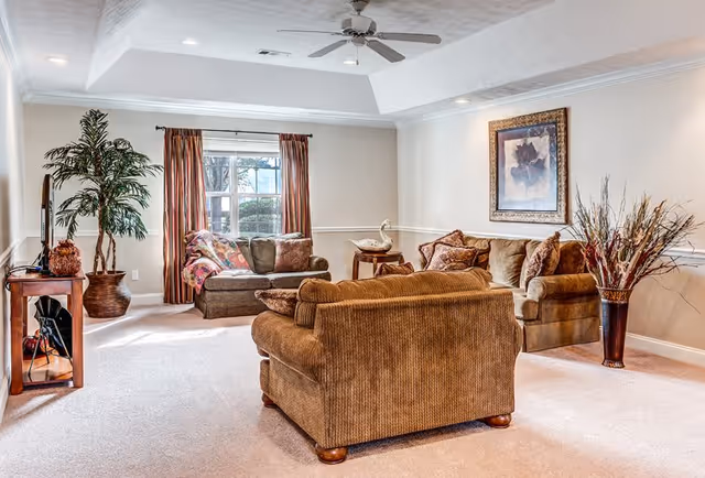 A cozy living room with beige carpet and light-colored walls featuring a ceiling fan. The room contains a brown upholstered sofa and loveseat with decorative pillows, a wooden side table with a white swan figurine, a large potted plant near a window with brown curtains, a framed floral artwork on the wall, and a tall vase with dried branches.
