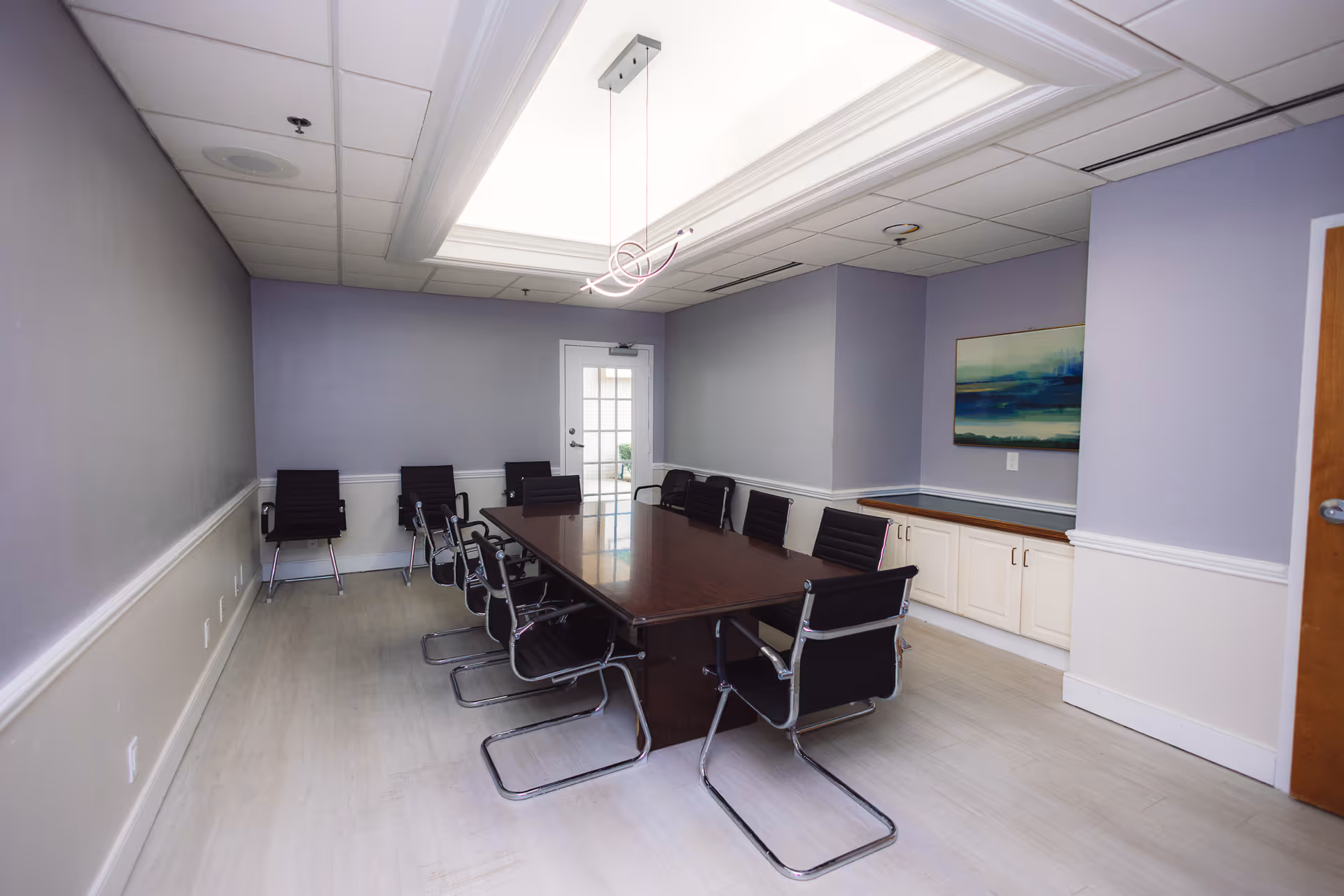 A conference room with a long dark wooden table surrounded by black office chairs. The walls are painted light purple with white trim, and there is a modern light fixture hanging from the ceiling. A door with glass panels is visible at the far end of the room, and a landscape painting hangs on the right wall above white cabinets.