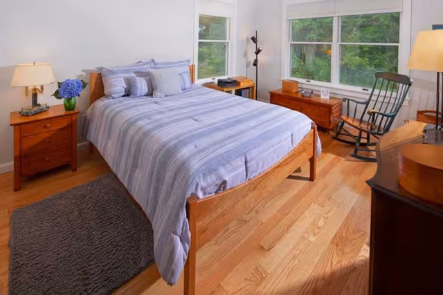 Bright bedroom with a wooden bed covered in blue-striped bedding, matching nightstands, a rocking chair and large windows overlooking greenery.