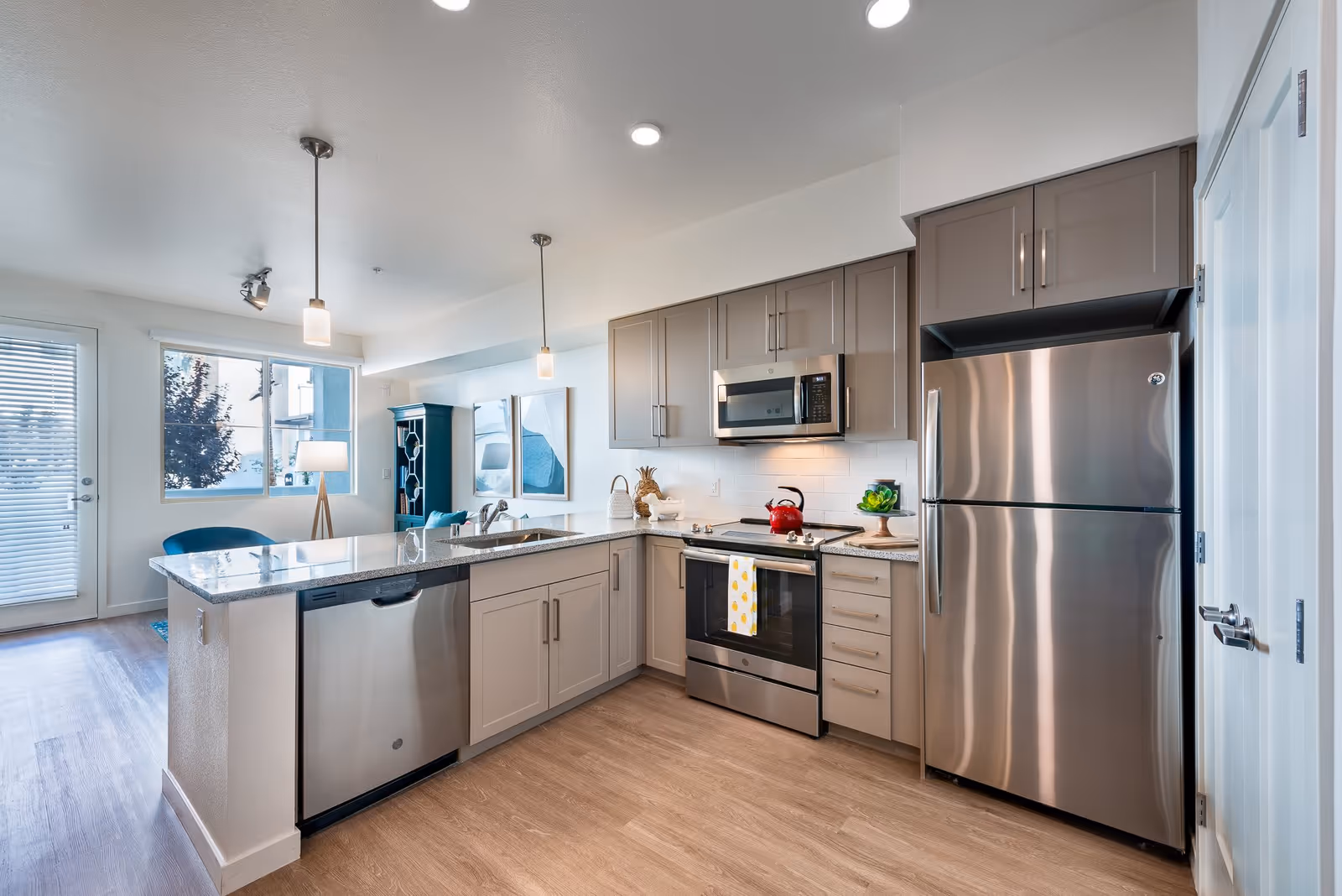 Modern kitchen with stainless steel appliances including a refrigerator, microwave, stove, and dishwasher. The kitchen features gray cabinets, a granite countertop island with a sink, two pendant lights hanging above, and a window and door leading outside. In the background, there is a blue chair, a floor lamp, and a cabinet.