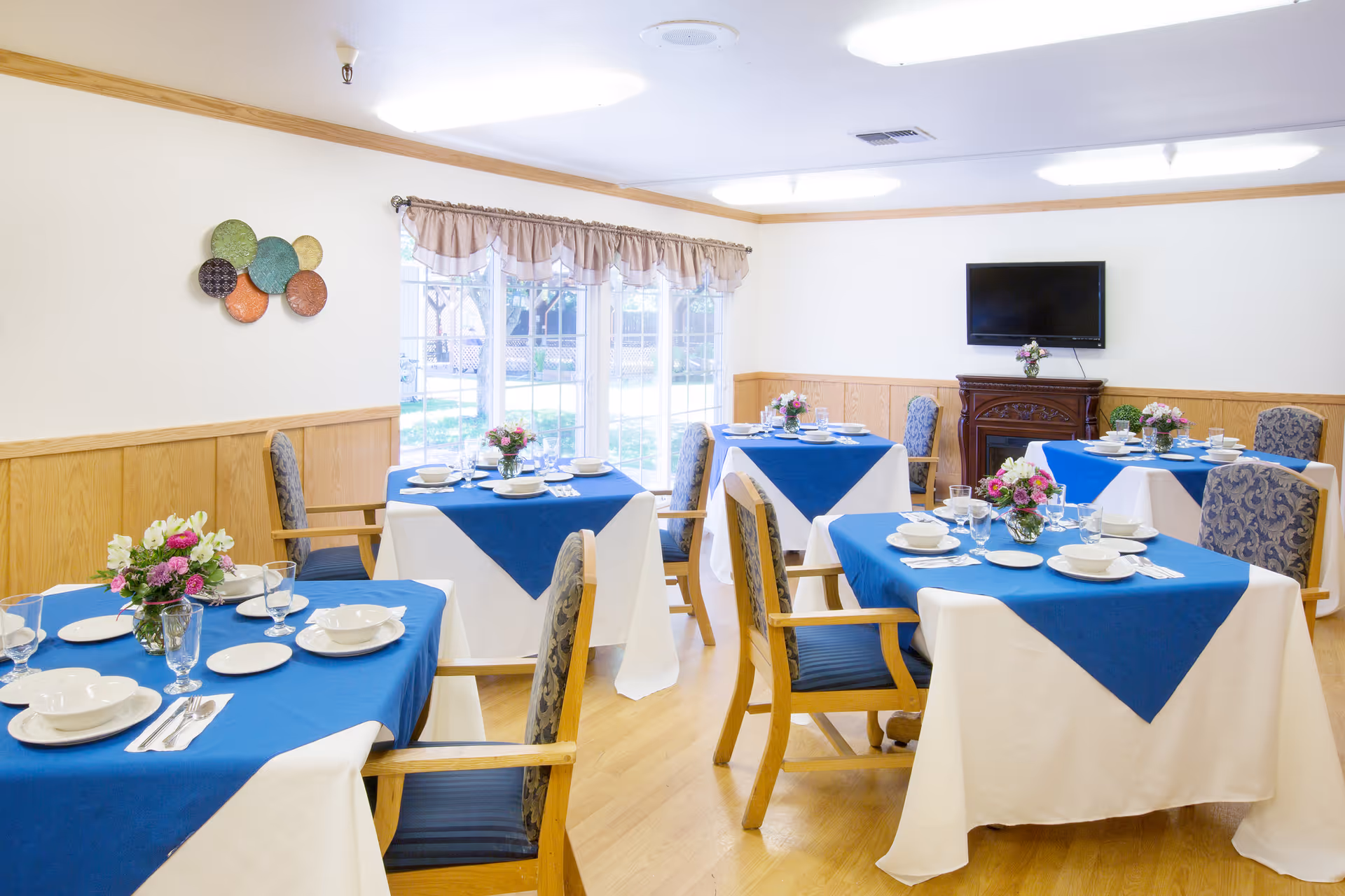 A bright dining room with four tables covered in white tablecloths and blue overlays, each set with plates, bowls, glasses, and silverware. Each table has a floral centerpiece. The room has wooden chairs with patterned cushions, wood paneling on the lower half of the walls, a wall-mounted TV above a decorative wooden cabinet, and large windows with sheer curtains letting in natural light.