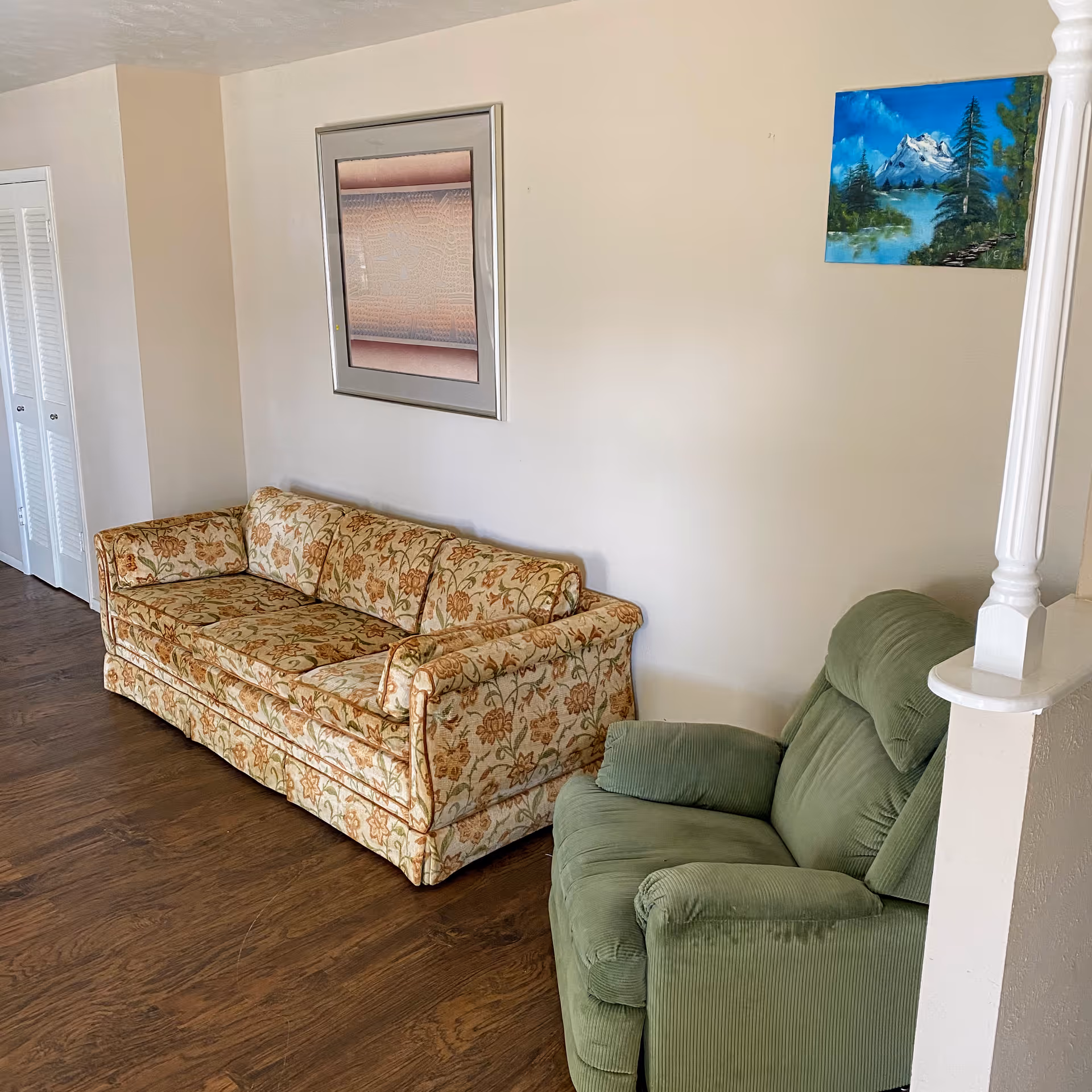 A living room area with a floral patterned sofa and a green upholstered armchair on a wooden floor. The walls are light-colored with two framed artworks hanging, one abstract and one landscape painting of mountains and trees.