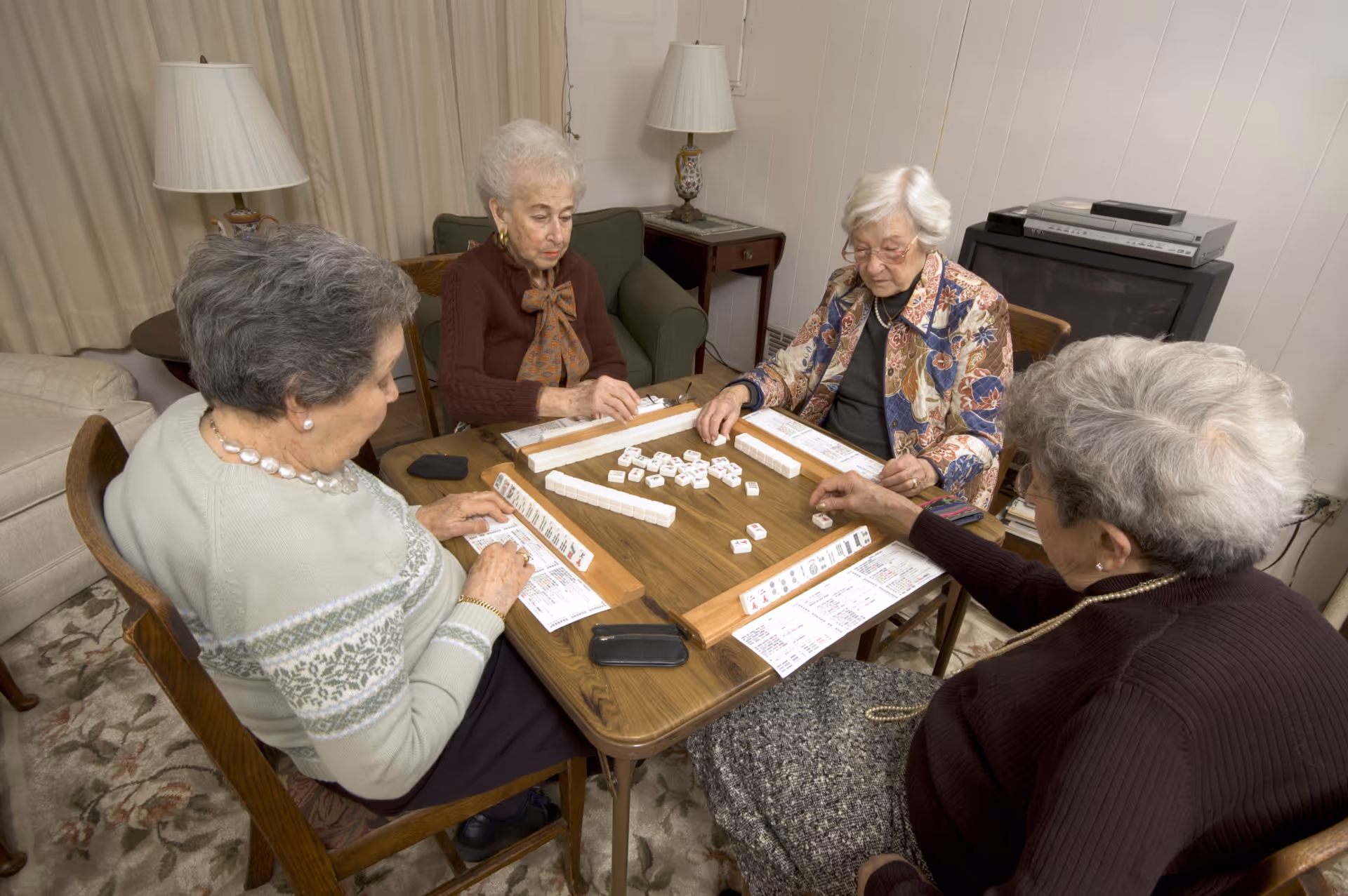 Four elderly women sitting around a wooden table playing a game with tiles in a cozy living room with a lamp, armchair, and television in the background.