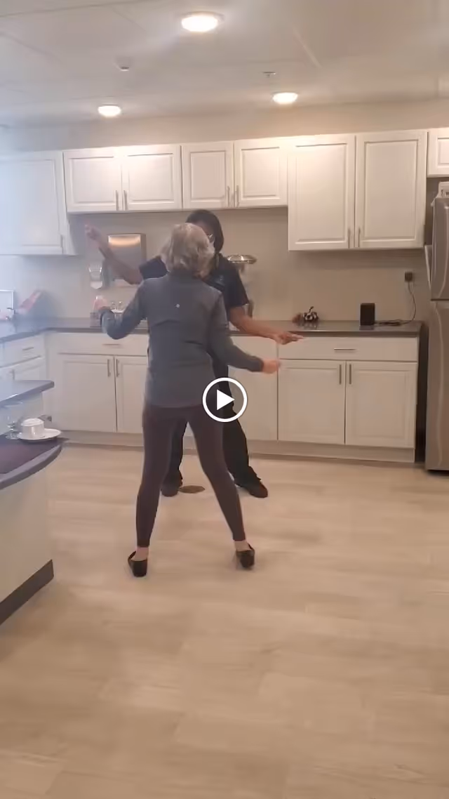 Two women dancing together in a modern kitchen with white cabinets, stainless steel refrigerator, and light wood flooring.