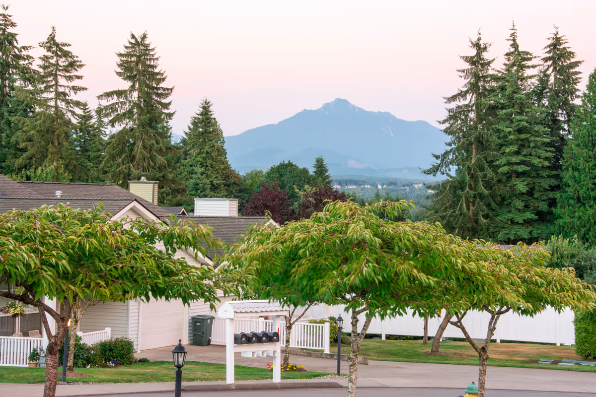 View of a senior living community with trees, rooftops of buildings, a mailbox cluster, and a mountain in the background under a clear sky at dusk.
