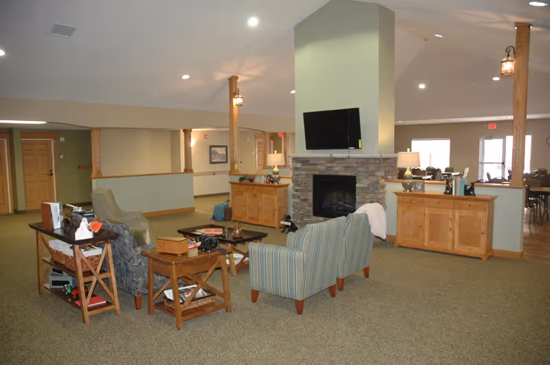 A spacious living room area in an assisted living facility featuring a stone fireplace with a mounted flat-screen TV above it. The room has several comfortable chairs and sofas arranged around a coffee table. Wooden cabinets flank the fireplace, and there are wooden support columns with hanging lantern-style lights. The room is carpeted and has neutral-colored walls with multiple doors and windows in the background.