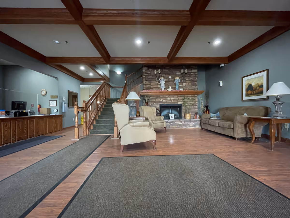 A cozy lobby-style interior with armchairs and a sofa facing a stone fireplace, a staircase to the upper level, and a reception desk under a wood-beamed ceiling.