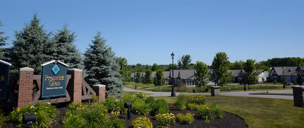 Entrance sign for Preswick Glen Distinctive Senior Living surrounded by landscaping with green bushes and yellow flowers, with a view of residential buildings and trees in the background under a clear blue sky.
