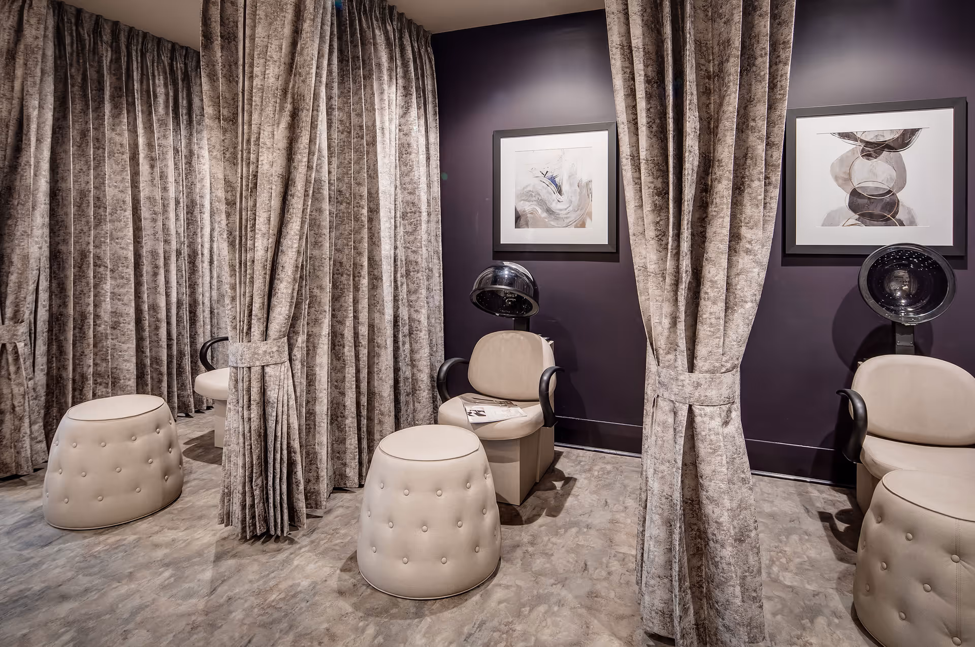 Interior view of a salon area with beige salon chairs and hair dryers, separated by thick beige curtains. The walls are painted dark purple and decorated with framed abstract artwork. The floor has a light-colored, textured finish.