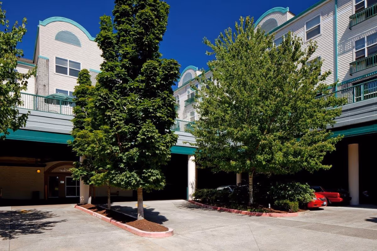 Exterior view of Hazelwood Community Apartments showing a multi-story building with white siding and teal trim. There are two large green trees planted in a landscaped area in front of the building, with a driveway and parked cars visible underneath a covered parking area. The sky is clear and blue.