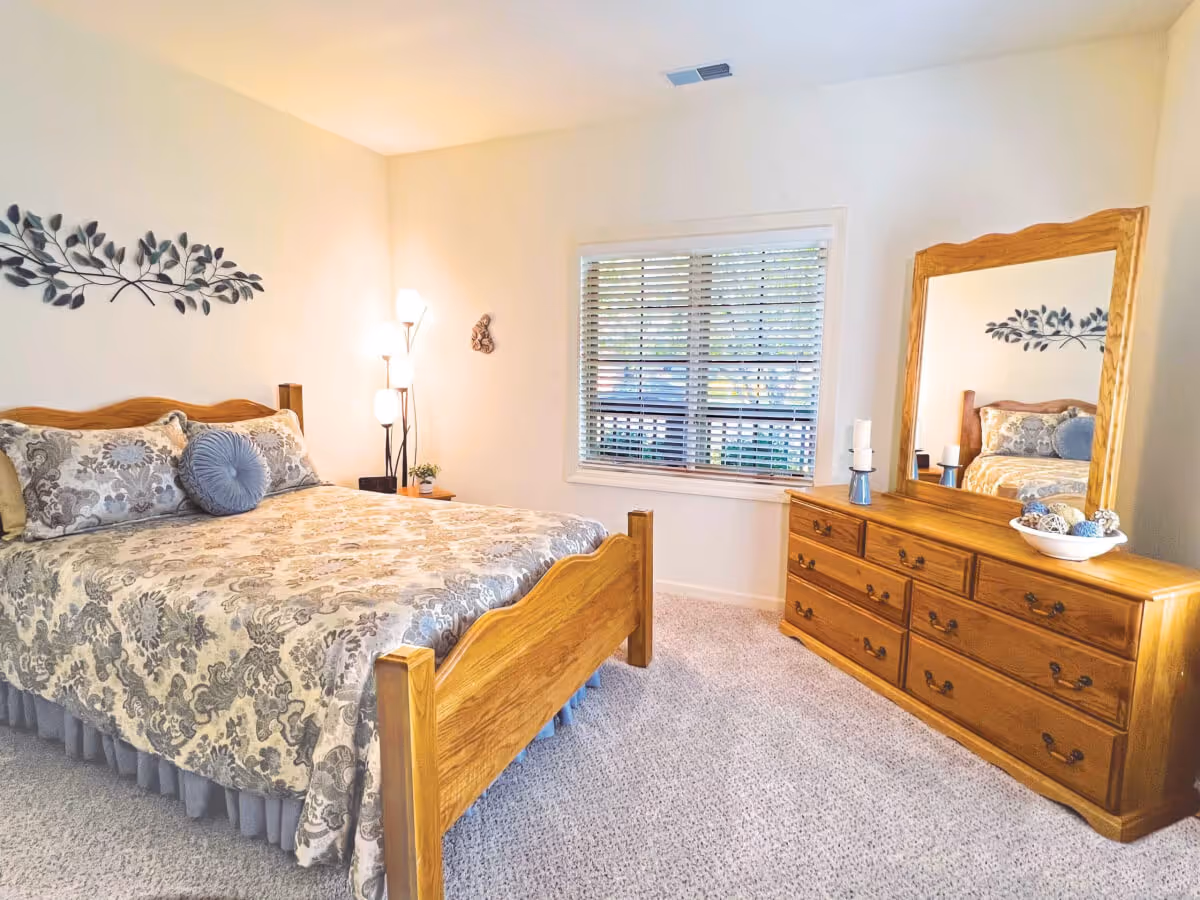 A cozy bedroom with a wooden bed frame and matching dresser with a large mirror. The bed is made with a patterned bedspread and decorative pillows. There is a window with blinds letting in natural light, a floor lamp with multiple lights, and a small plant on a nightstand. The walls are light-colored and there is a decorative metal wall art above the bed.