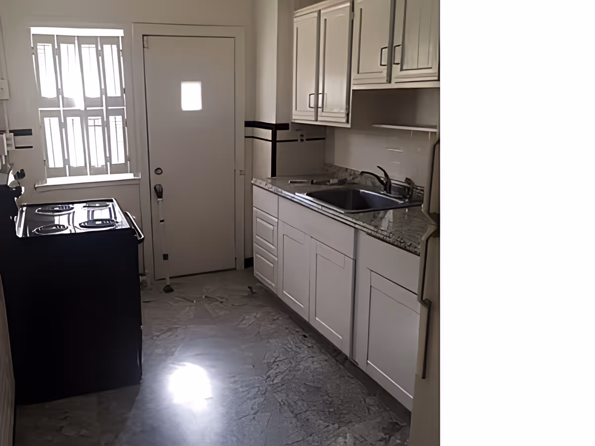 Narrow kitchen with white cabinets and granite countertop with a sink on the right and a stove on the left near a door and window.