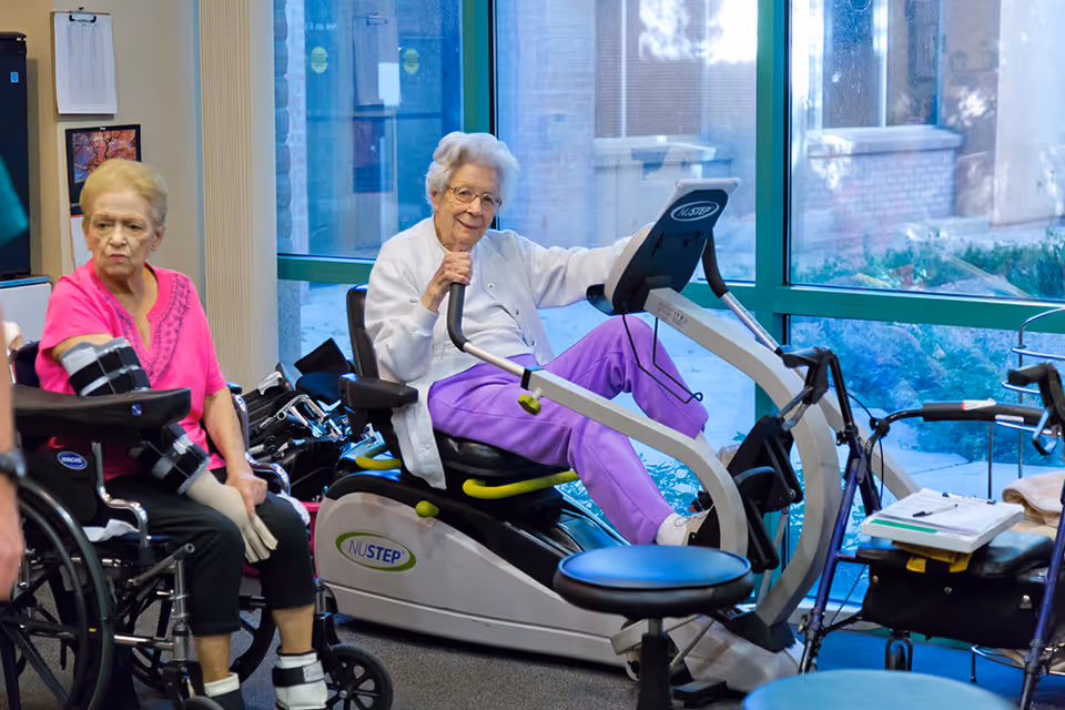 Two elderly women in a senior living facility exercise room. One woman is seated on a NuStep exercise machine, smiling and wearing a white jacket and purple pants. The other woman is sitting in a wheelchair, wearing a pink top and a black arm brace. Large windows show an outdoor view with plants and a brick building.