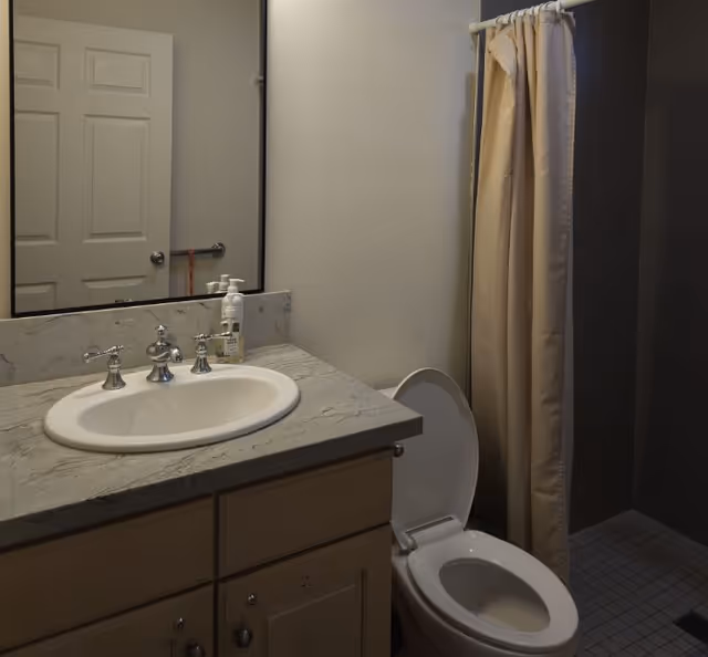 A bathroom with a white sink set in a marble countertop, a large mirror above the sink, a toilet with the lid open, and a shower area with a beige curtain.