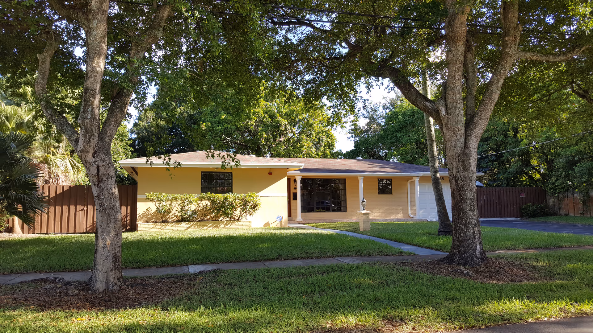 Single-story yellow house with a brown roof, a front lawn with green grass, two large trees in the foreground, a driveway on the right side, and a wooden fence surrounding the property.