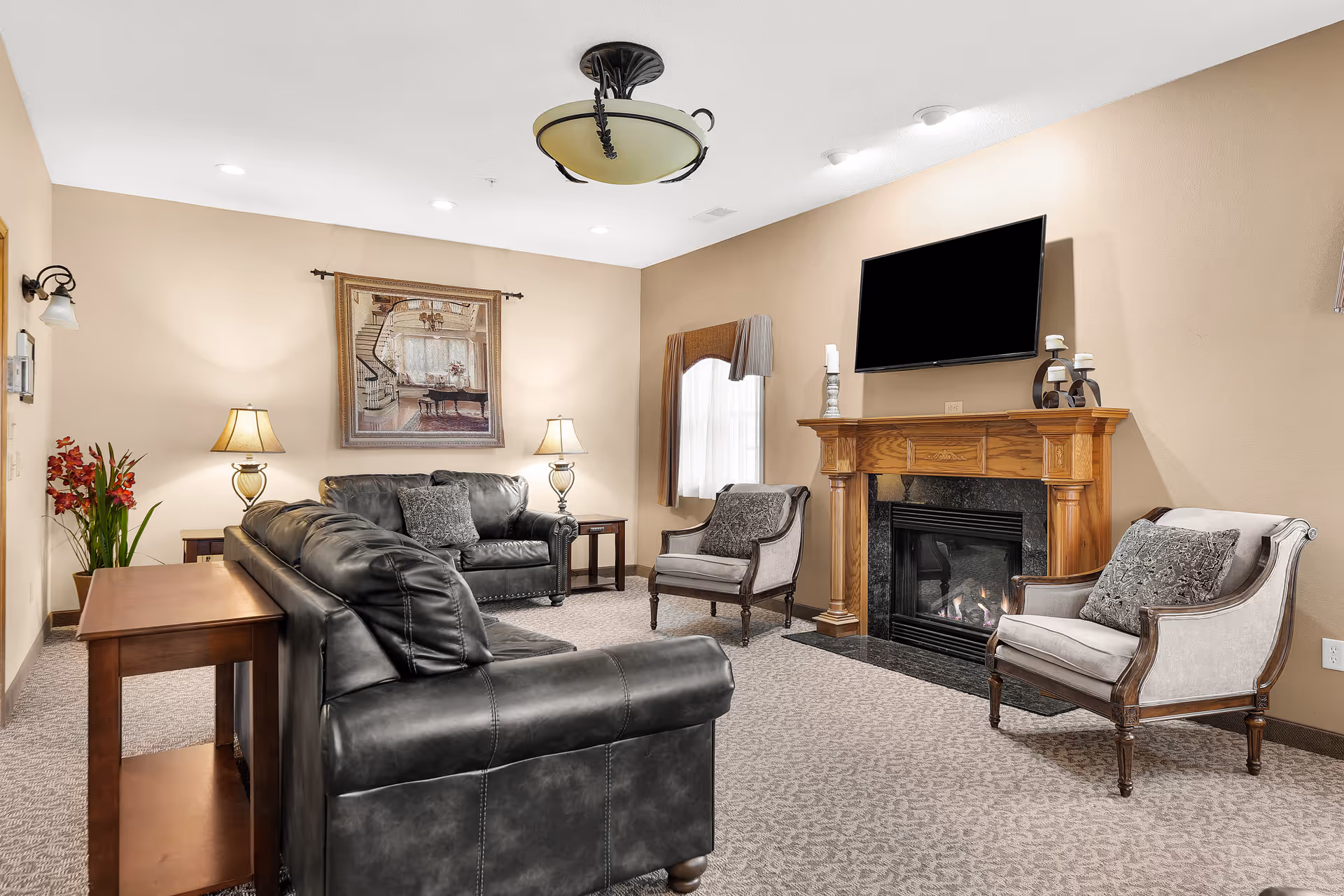 A cozy living room with beige walls and carpeted floor featuring two black leather sofas, two upholstered armchairs with decorative pillows, a wooden fireplace with a mounted flat-screen TV above it, two table lamps on side tables, a framed artwork on the wall, and a window with a valance curtain.