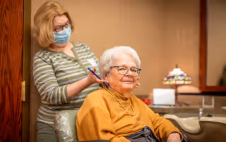 An elderly woman with white hair and glasses sits in a chair smiling while a caregiver wearing a face mask stands behind her, writing on a clipboard in a warmly lit room with wooden walls and a decorative lamp in the background.