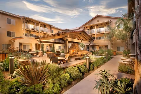 Outdoor courtyard with a covered seating area, chairs and tables, landscaped pathways, and a surrounding three-story building with balconies.