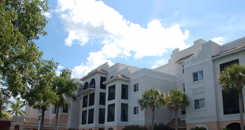 Front exterior of a multi-story white residential building with screened balconies, palm trees, and a blue sky.